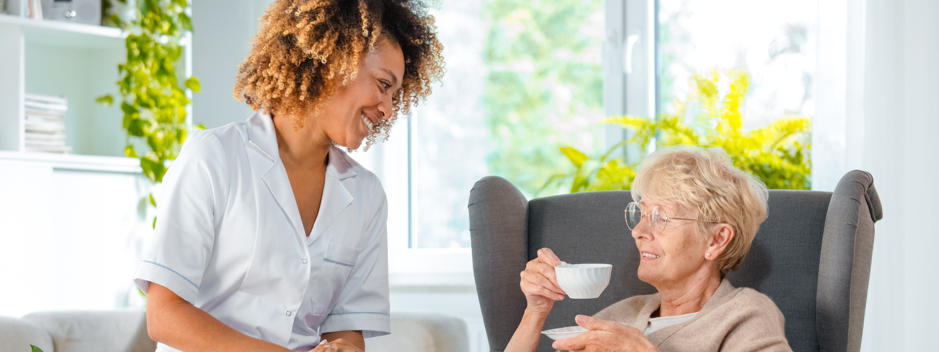 A carer smiling at a care home resident sitting in a chair having a cup of tea.